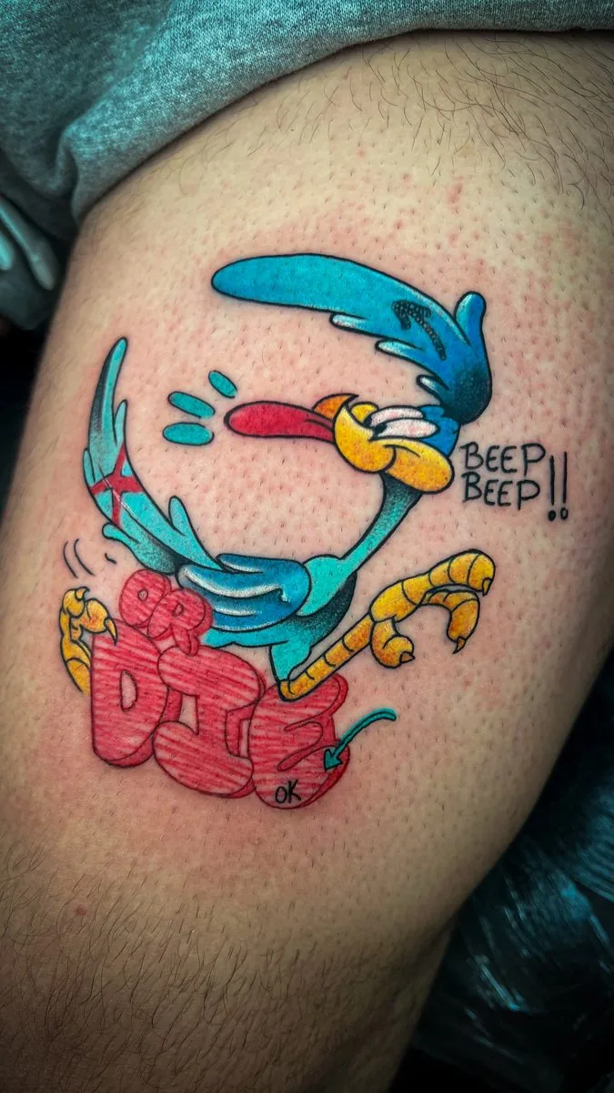 Tatuaje estilo parche bordado (patch): pájaro de caricatura corriendo sobre letras rojas, con textura de hilo, en la pierna