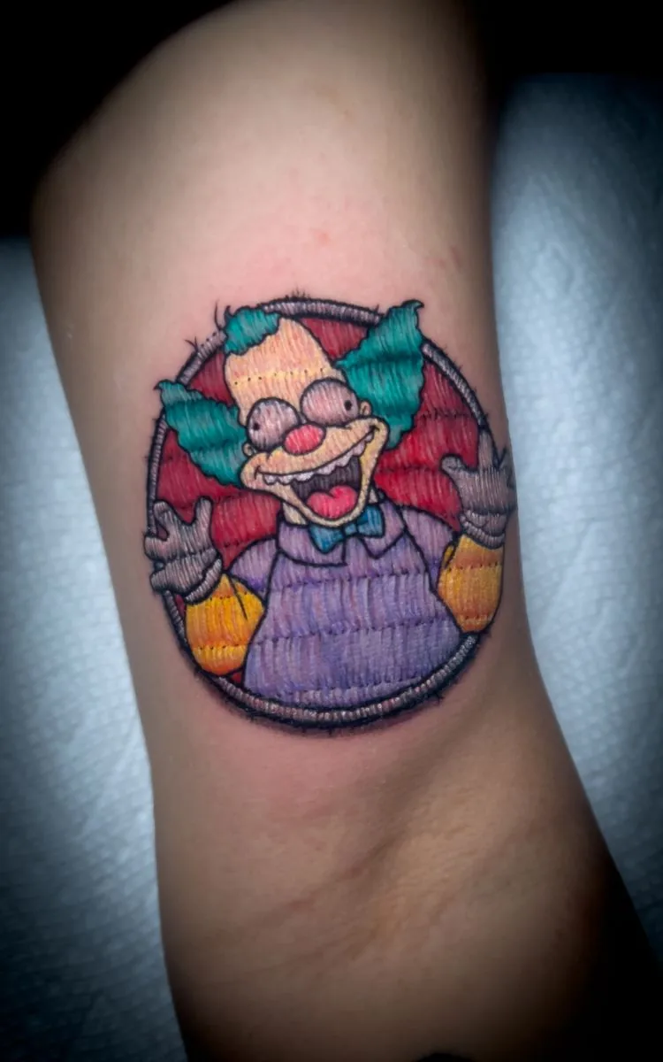 Tatuaje estilo parche bordado: payaso de caricatura en un parche redondo, en el brazo