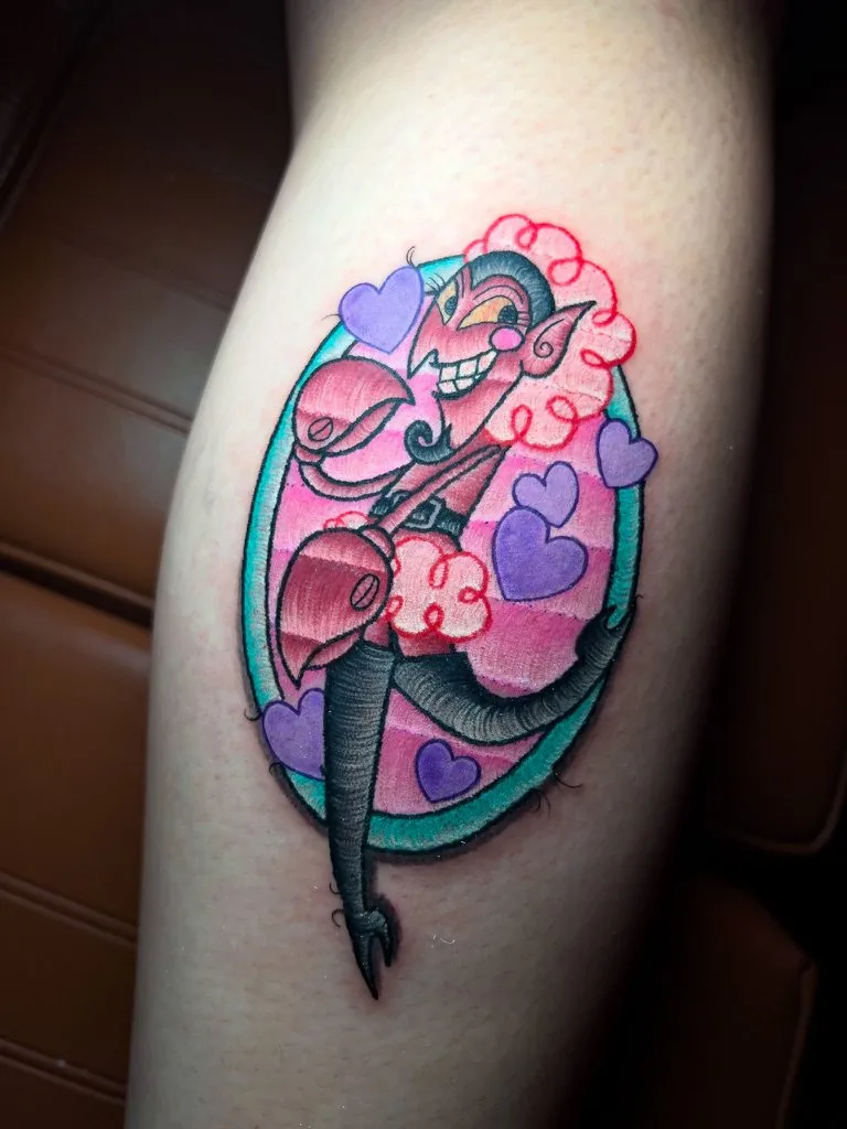 Tatuaje a color estilo parche: personaje de caricatura con corazones en un óvalo con borde bordado, en la pierna